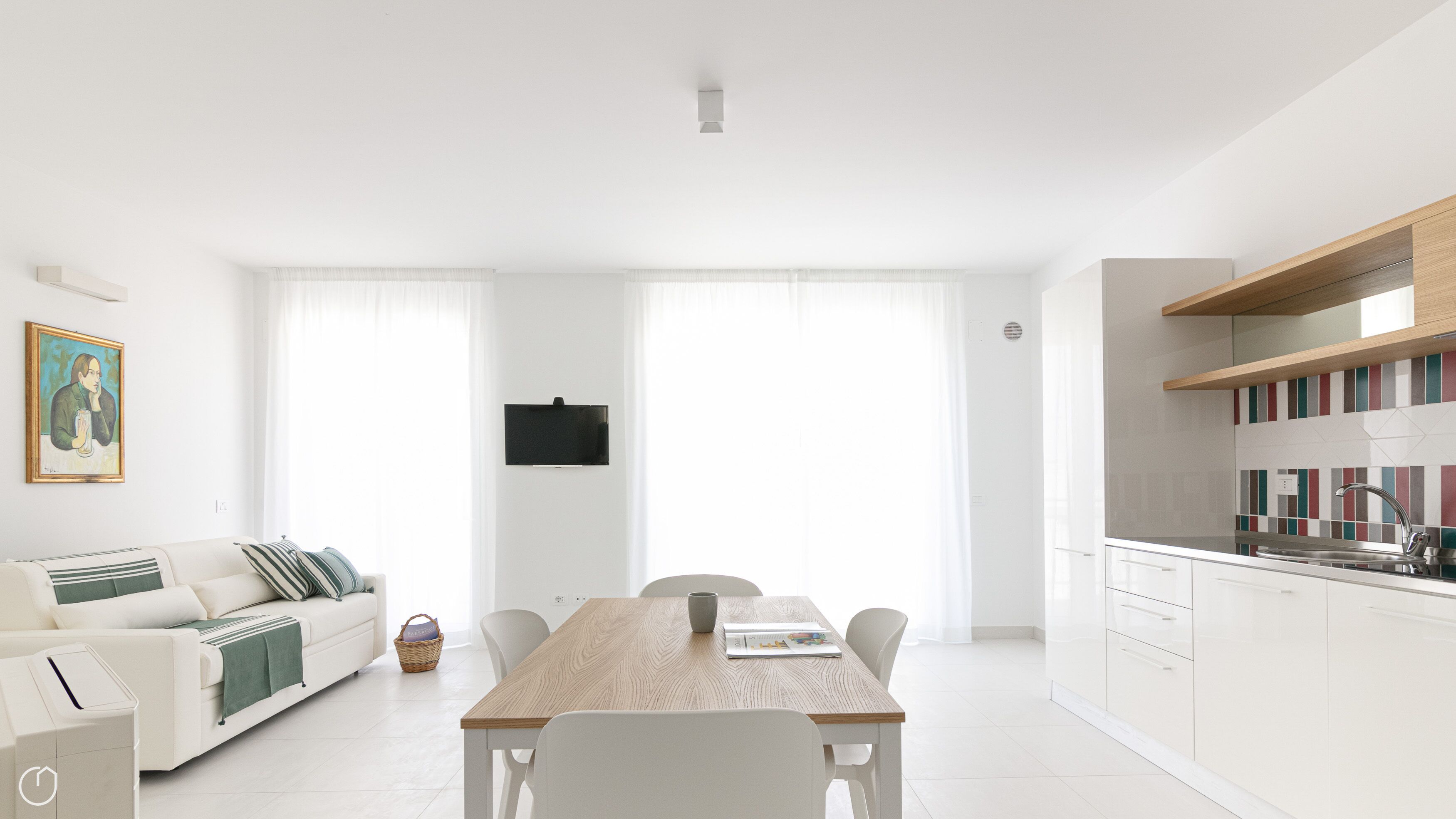 Un salon moderne avec un canapé blanc, une table à manger et un coin cuisine doté de meubles blancs et d’une étagère en bois.