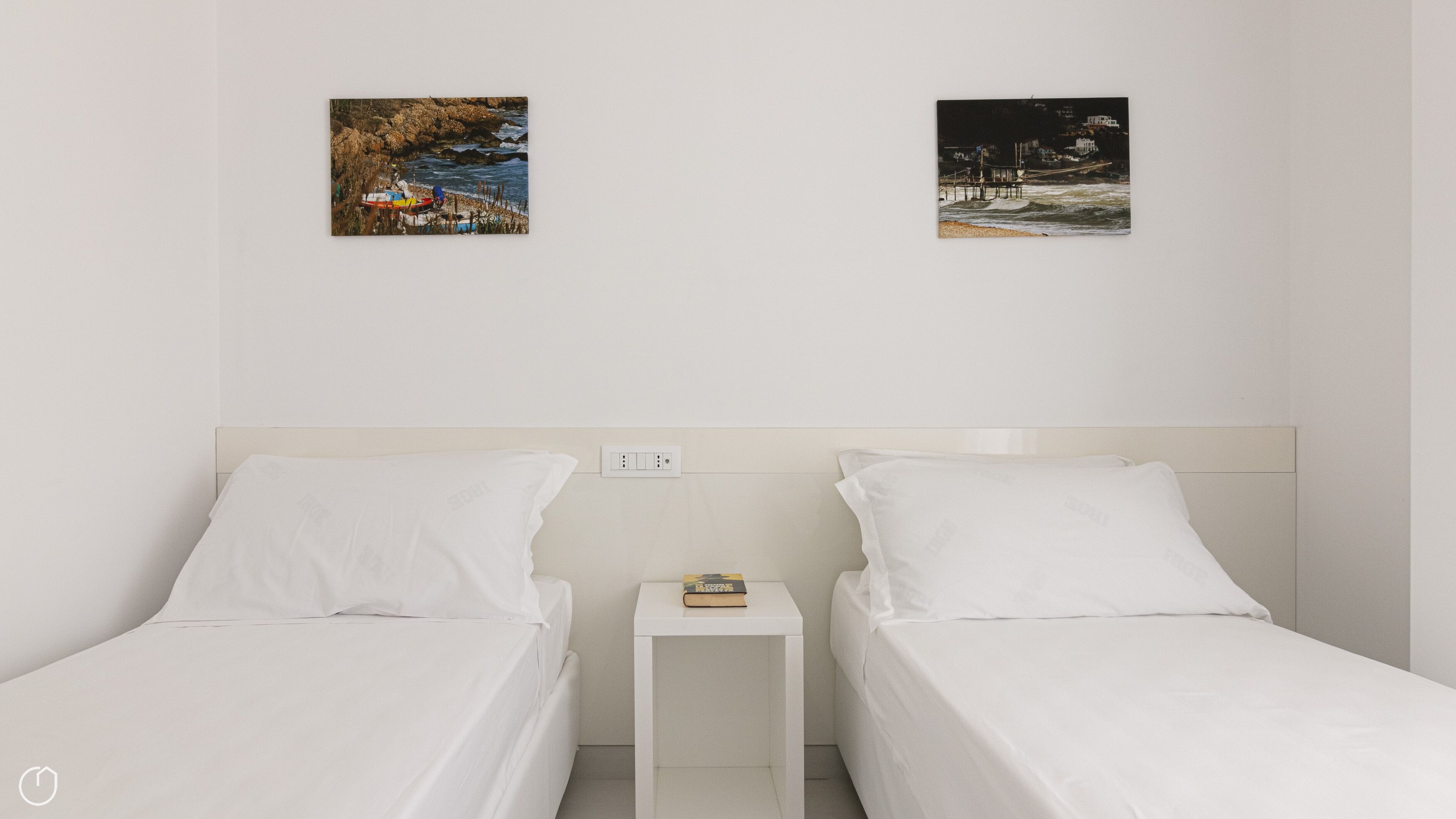 Une chambre d’hôtel avec deux lits simples, une petite table de chevet sur laquelle se trouve un livre, et deux tableaux encadrés accrochés au mur.