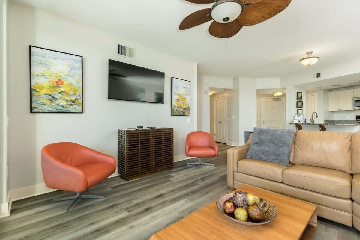 Een woonkamer met een beige bank, twee oranje fauteuils, een houten salontafel met fruit, een plafondventilator en een flatscreen televisie aan de muur.