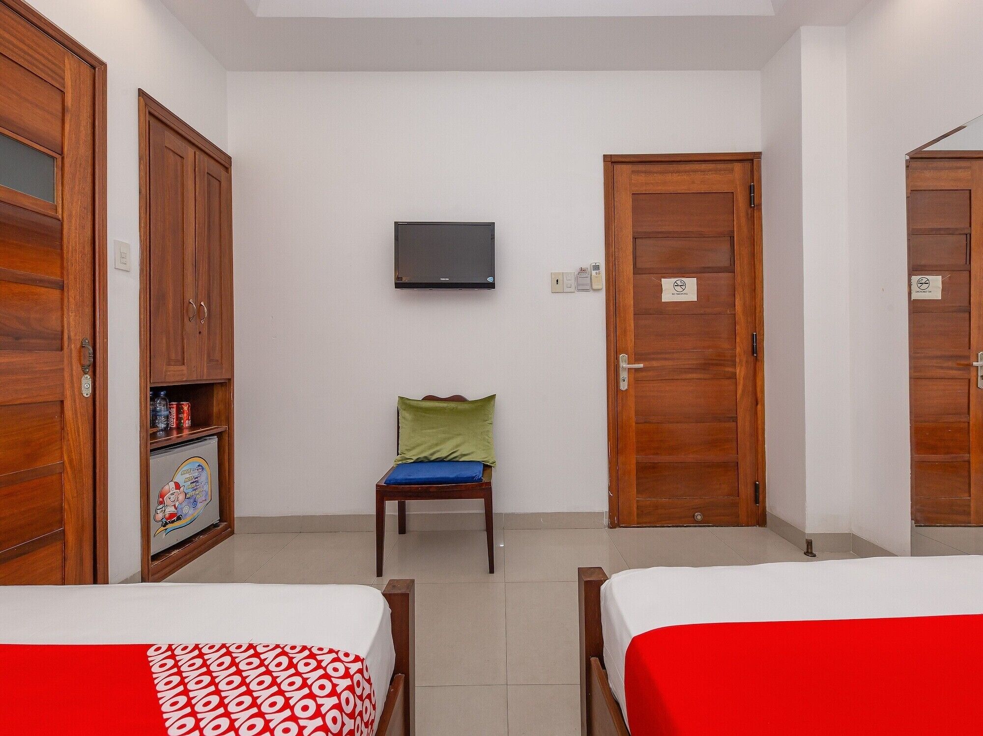 A hotel room with two beds, a television, wooden wardrobes, and a small table with a green cushion.