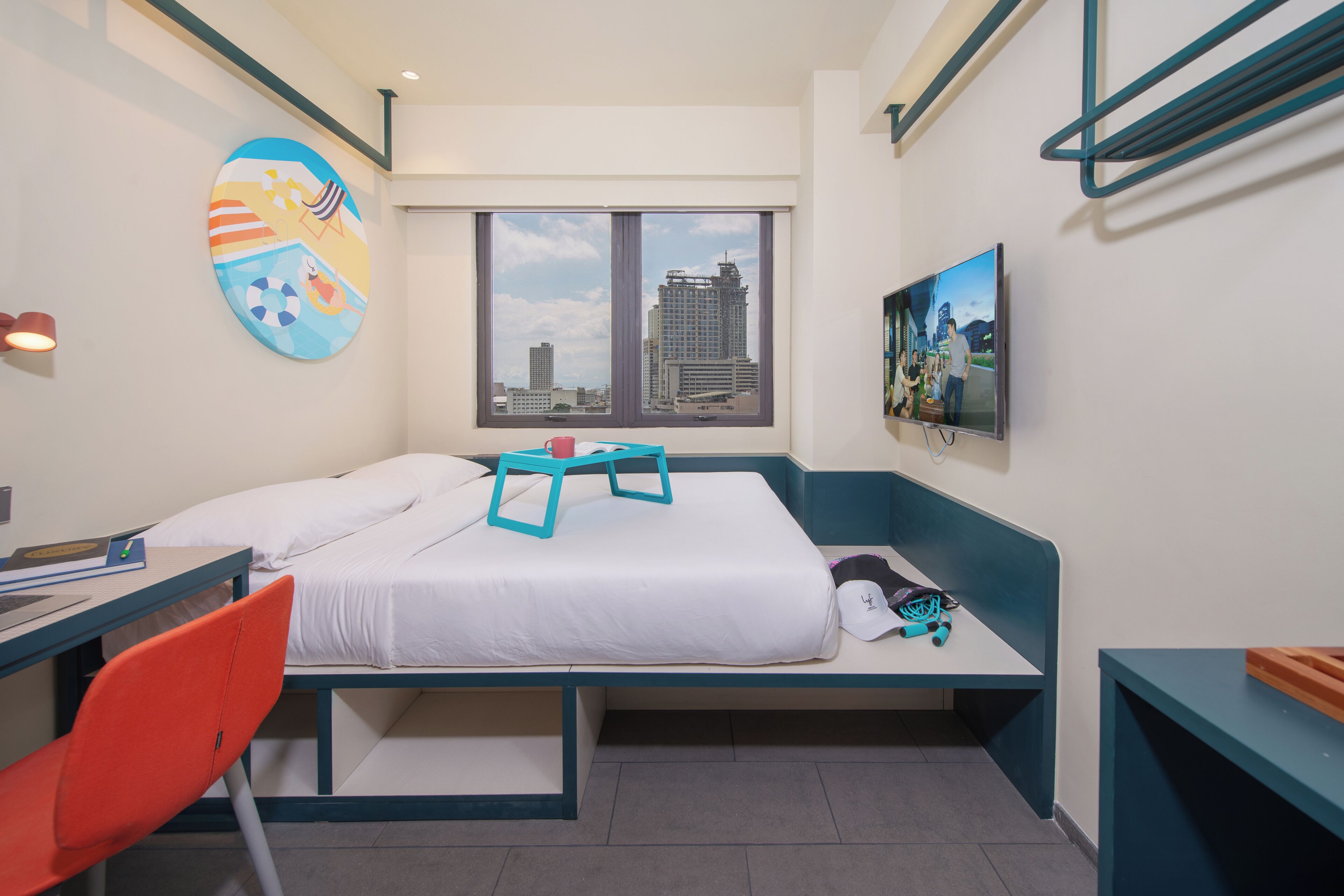 A hotel room with a bed, a desk, a chair, a window with a city view, and a colorful wall decoration.