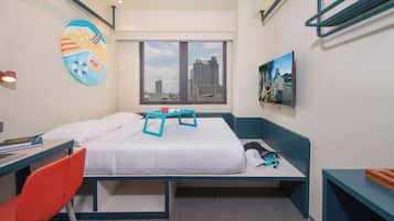 A hotel room with a bed, a desk, a chair, a window with a city view, and a colorful wall decoration.