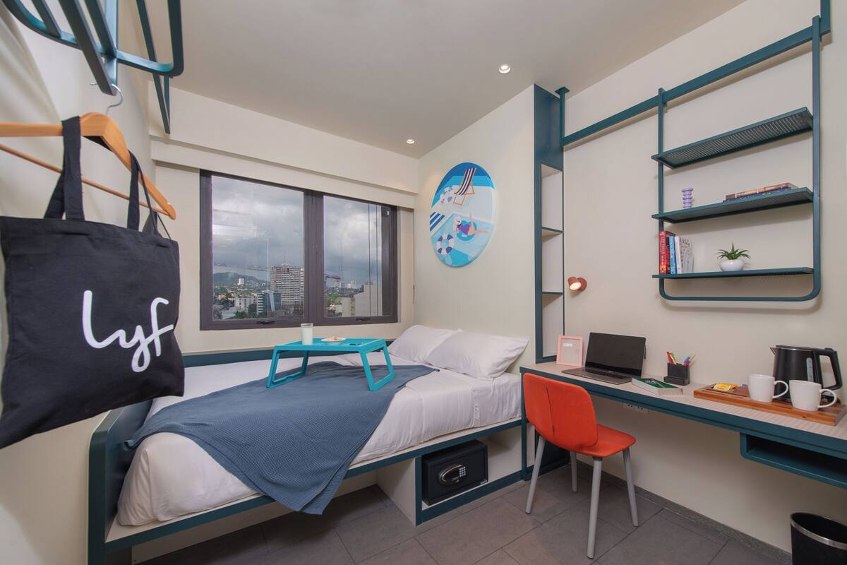 Quarto de hotel pequeno com uma cama, uma escrivaninha, uma cadeira, um aparelho de café e vista para a cidade pela janela.