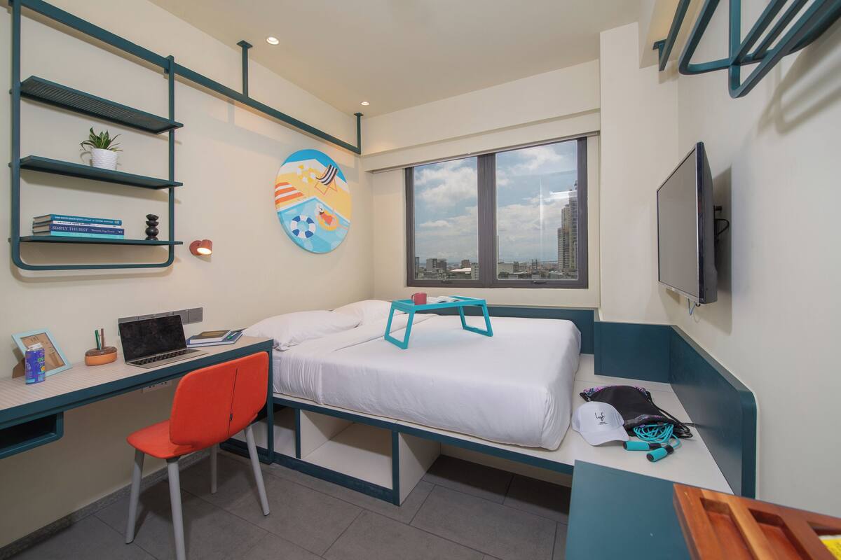 Quarto de hotel compacto com cama, escrivaninha com laptop, cadeira, televisão e janela com vista para a cidade.