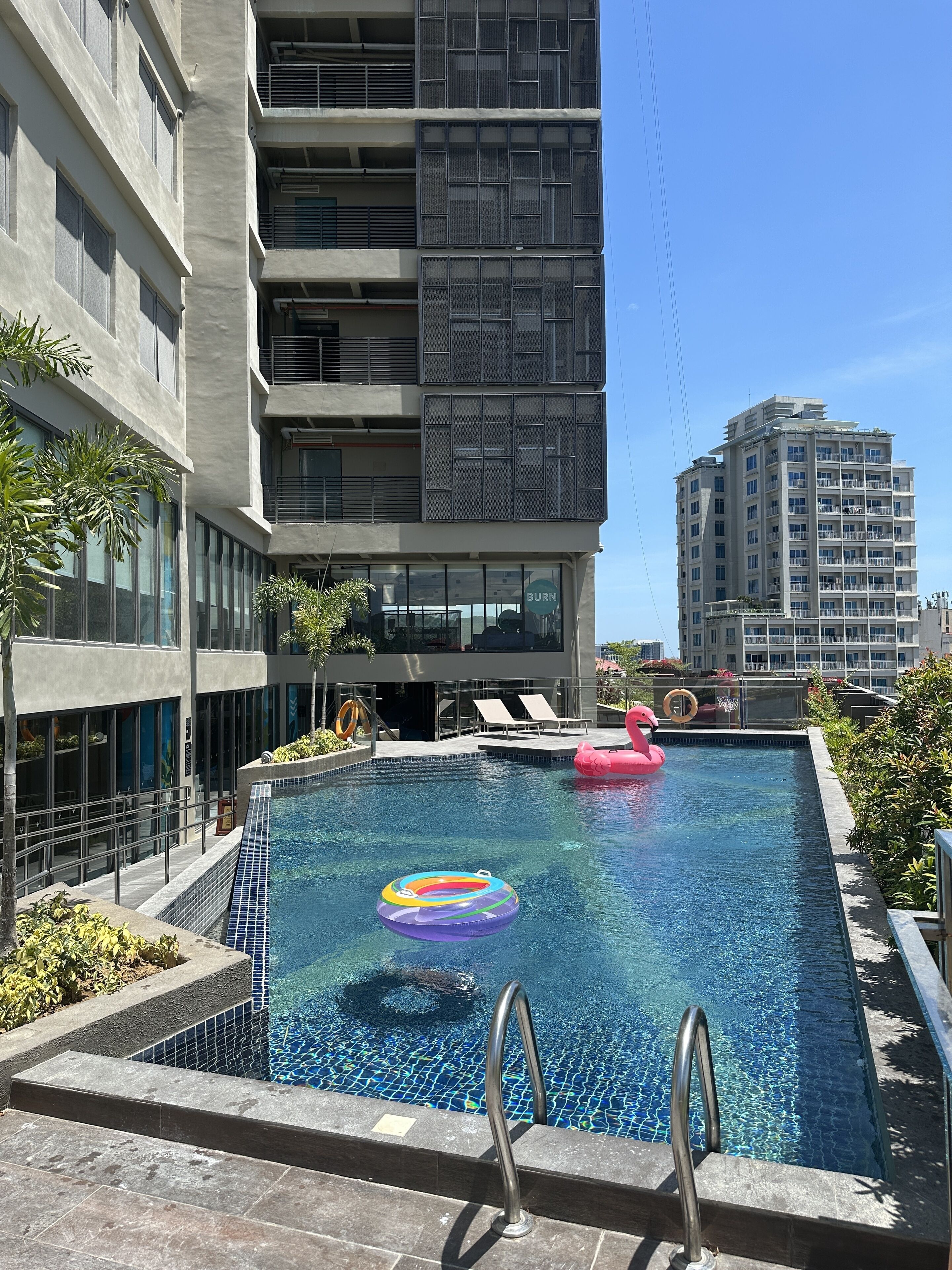 Un edificio moderno con área de piscina en la azotea, que incluye una piscina, sillas de descanso y un flotador con forma de flamenco rosa.