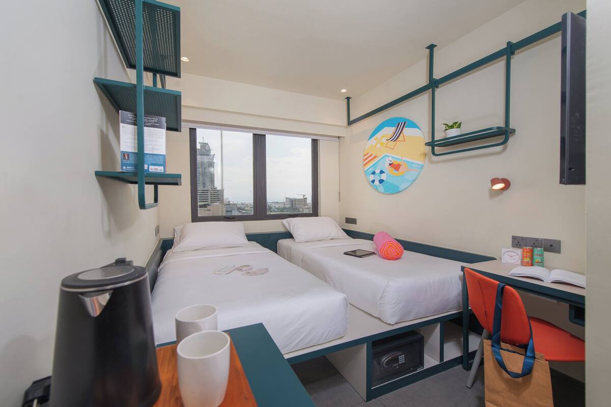 Quarto de hotel com duas camas, uma escrivaninha com cadeira, cafeteira, lixeira e janela com vista para a cidade.