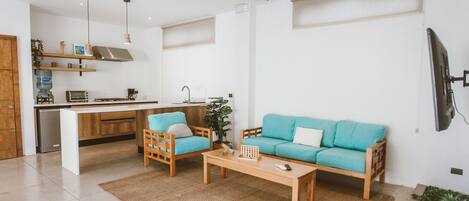 Ein modernes Wohnzimmer mit Sofa, Couchtisch und Sesseln. Es gibt eine Küchenzeile mit Herd, Spüle und Regalen. Ein Deckenventilator und ein Fernseher sind an der Wand befestigt.