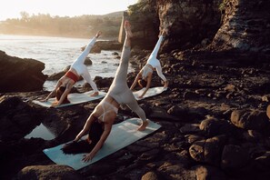 Three individuals performing yoga on mats on a rocky beach with a cliff in the background.