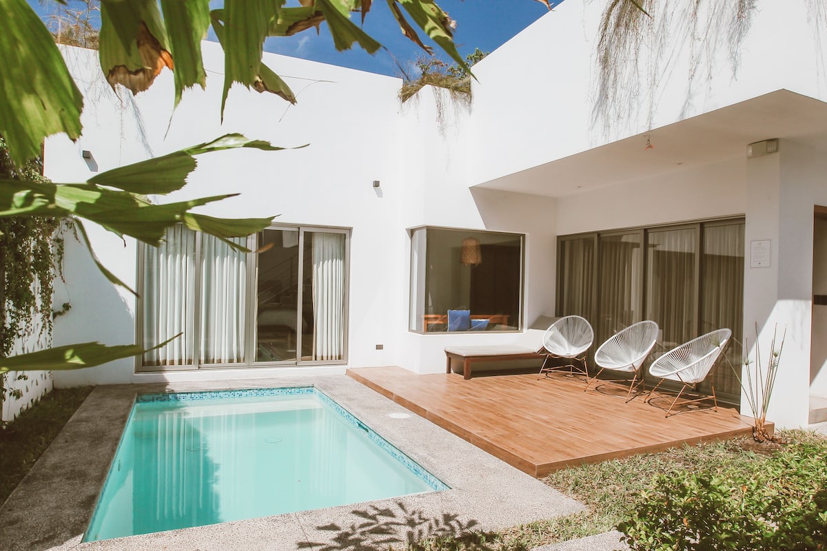 A modern outdoor pool area with a wooden deck, white chairs, and a glass door leading to a covered patio.