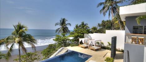 A modern pool area with a view of the beach and ocean, a sunbed, and a canopy bed.