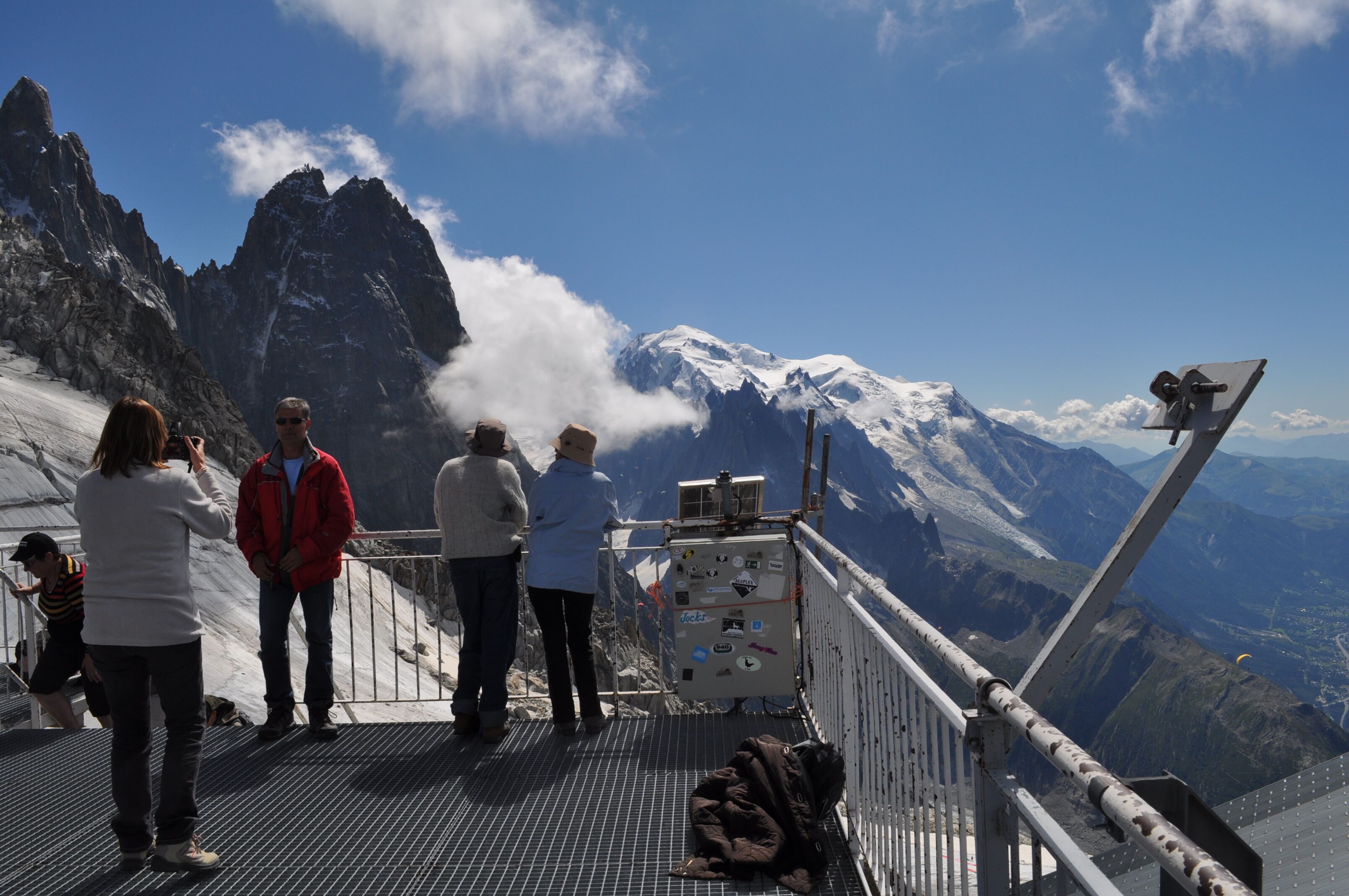 A group of people on a metal platform overlooking a mountainous landscape with snow-capped peaks and a clear sky.