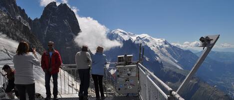 A group of people on a metal platform overlooking a mountainous landscape with snow-capped peaks and a clear sky.