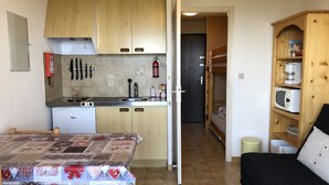 Una cocina compacta con armarios de madera, un lavamanos y un microondas. Hay una mesa de comedor con mantel estampado y una cama nido con colchón blanco y negro.
