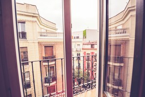 A view through a window with a balcony railing, overlooking a street with buildings of varying heights and architectural styles.