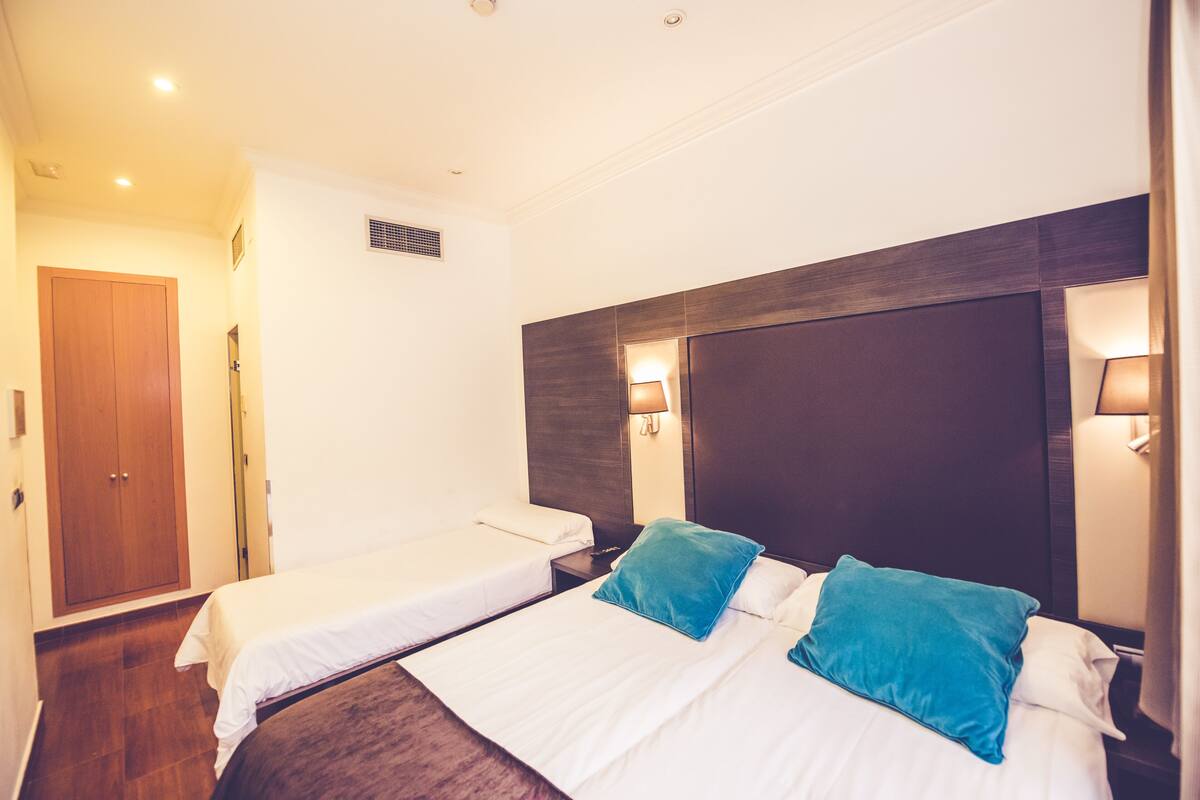 Habitación de hotel con dos camas individuales, un armario de madera, una mesita de noche y lámparas de pared.