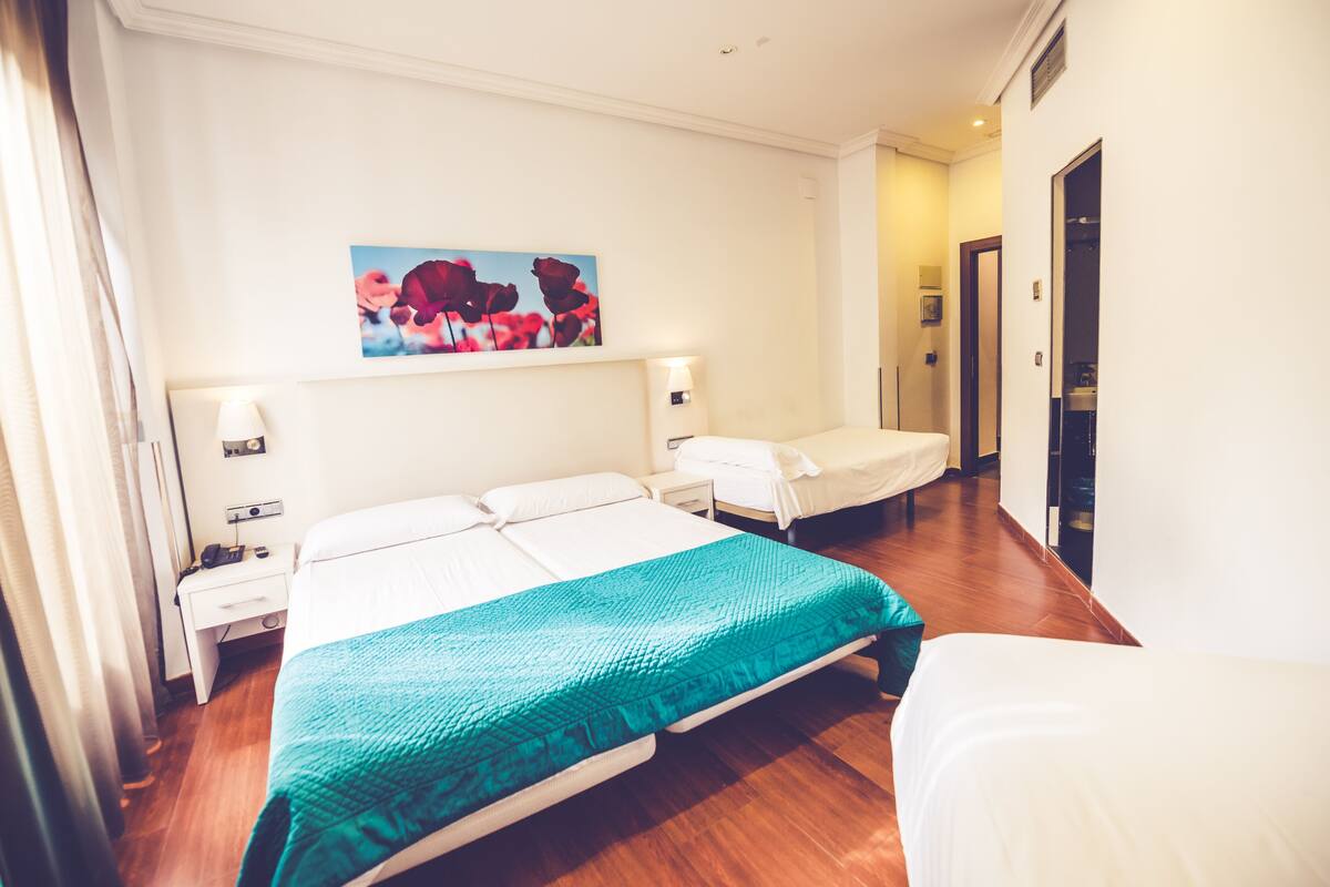 Un cuarto de hotel con una cama grande, una cama más pequeña, una mesita de noche, una lámpara, un cuadro en la pared y piso de madera.