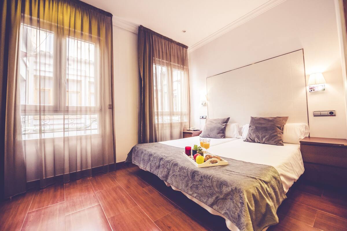 Una habitación de hotel con una cama grande, una mesita de noche, cortinas en la ventana y una lámpara de pared.