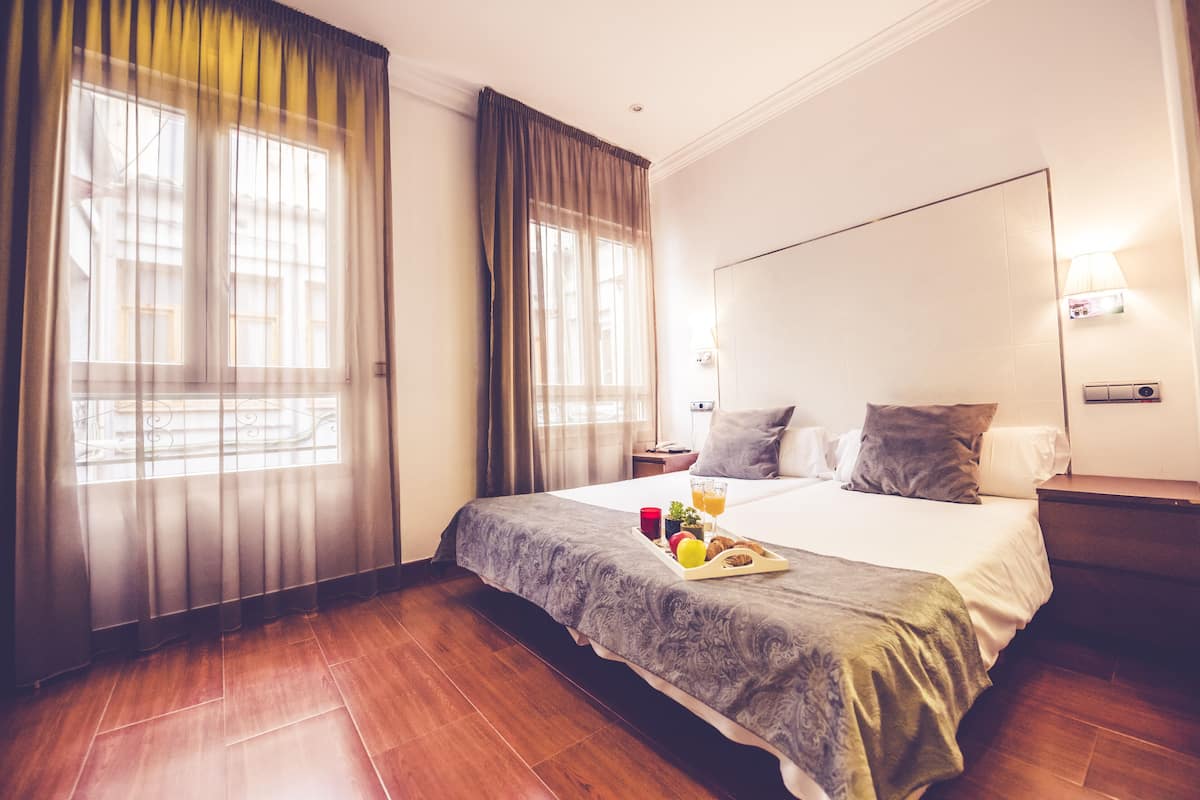 A hotel room with a large bed, a bedside table, a nightstand, a window with curtains, and a wall-mounted lamp.