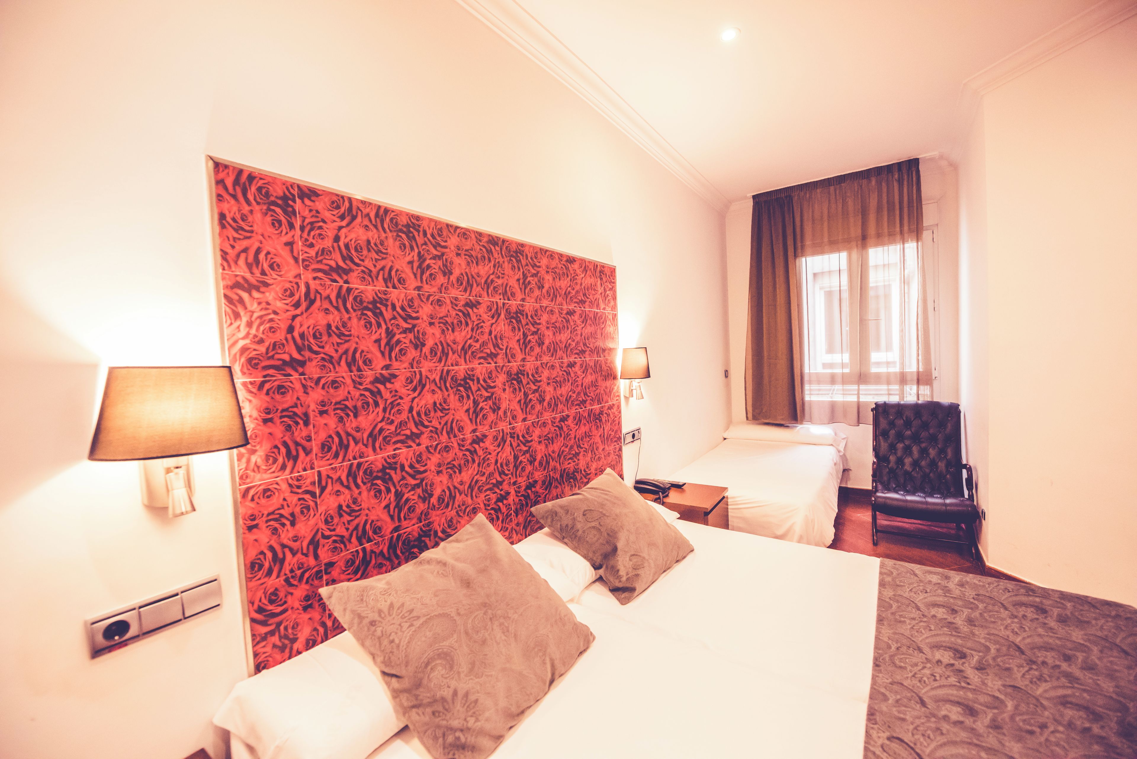 A hotel room with two beds, a red patterned headboard, a chair, and a window with curtains.