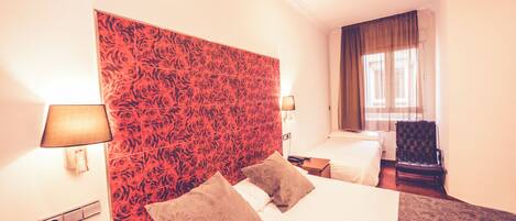 Une chambre d’hôtel avec deux lits, une tête de lit rouge à motifs, une chaise et une fenêtre avec des rideaux.
