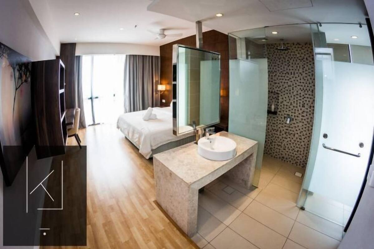 A modern hotel room with a bed, a large mirror, a bathroom with a glass shower enclosure, and a wooden floor.