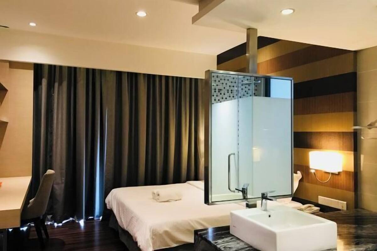 A hotel room with a bed, a bathroom with a glass shower enclosure, a marble countertop, and a wall-mounted light fixture.
