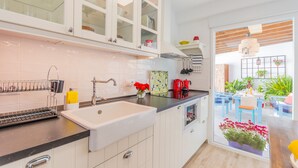 Una cocina moderna con armarios blancos, encimera negra y un lavamanos. Se observa un comedor a través de una puerta de vidrio con sillas azules y una mesa.