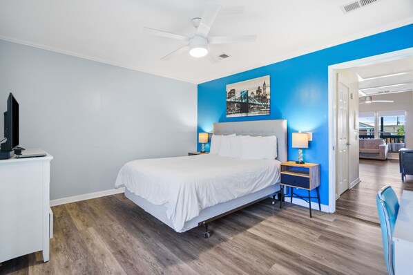A hotel room with a bed, a desk, a chair, a ceiling fan, and a painting on the wall.