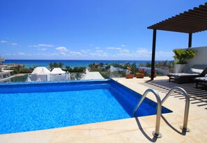 A swimming pool with a view of the ocean, a covered patio with lounge chairs, and a pergola with potted plants.