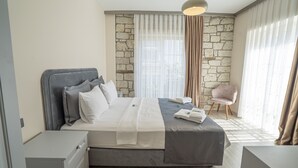 Ein modernes Schlafzimmer mit einer Wand aus Stein, einem großen Bett mit grauer Bettwäsche, einem Nachttisch, einem Sessel und einem Fenster mit durchsichtigen Vorhängen.