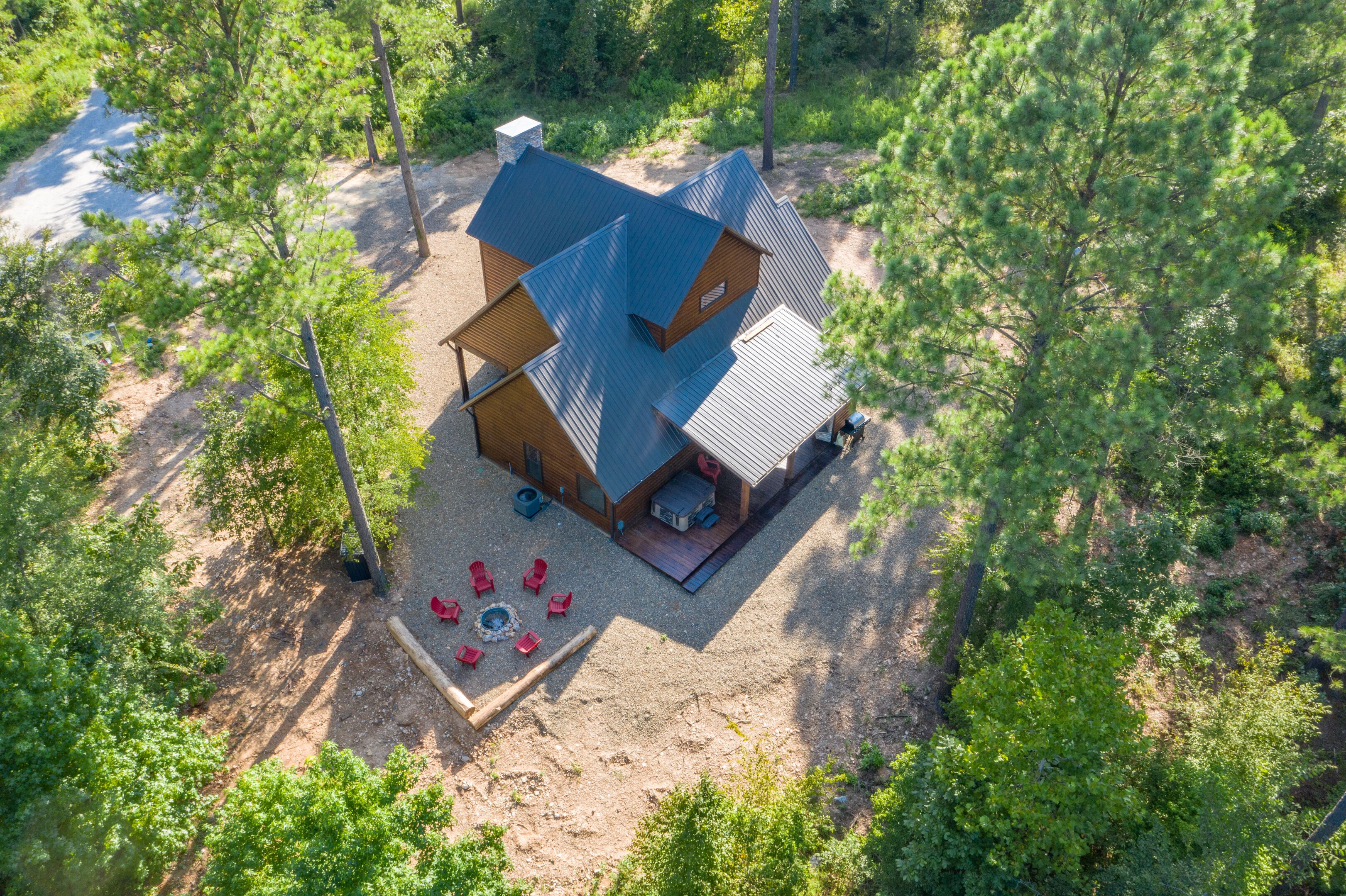 A cabin with a sloped roof, surrounded by trees, and a fire pit with red chairs.