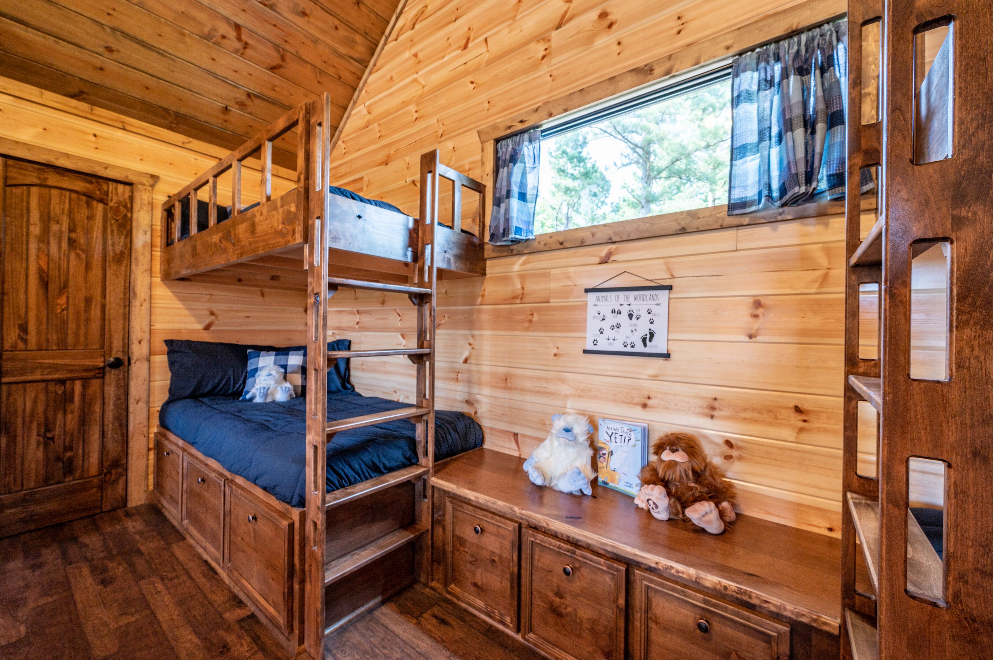 Une pièce de cabane en bois avec des lits superposés, une fenêtre avec des rideaux, un calendrier accroché au mur et une petite table avec des jouets.