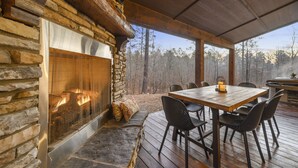 Une cheminée en pierre avec un feu de bois, une terrasse en bois avec une table et des chaises, et une vue sur les arbres à travers de grandes fenêtres.