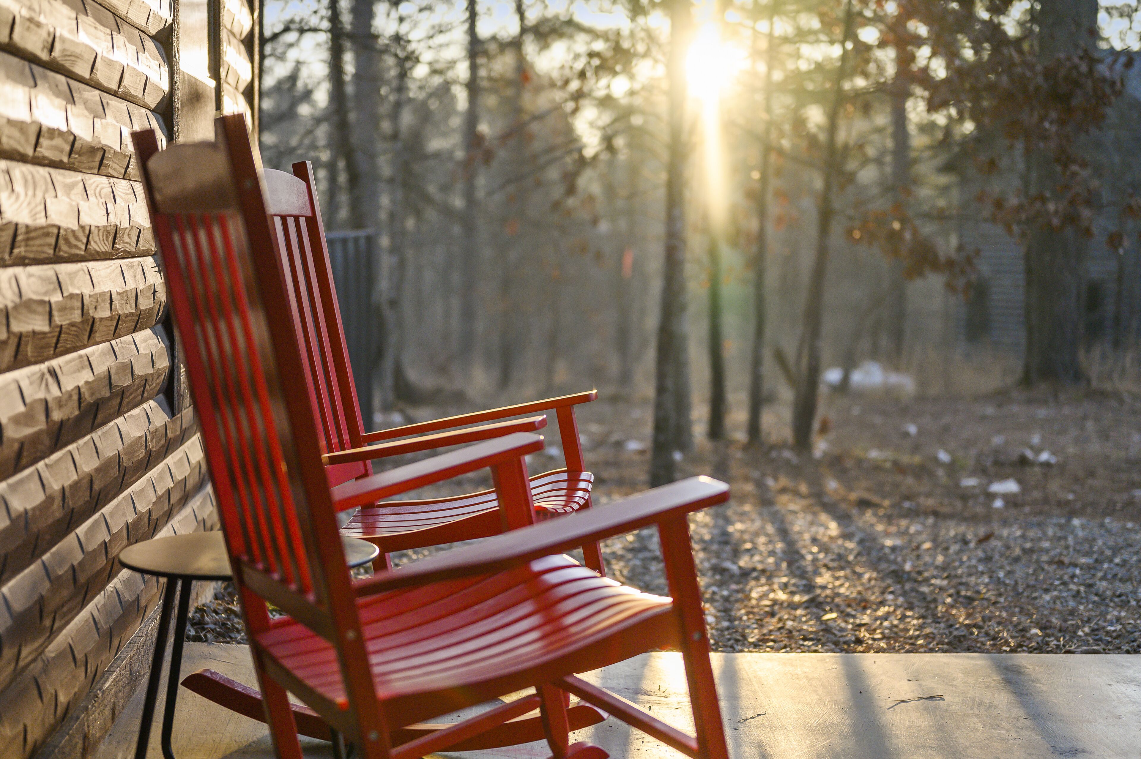 A wooden chair with a high backrest and armrests, positioned outdoors with a brick wall to the left and a forested area in the background.
