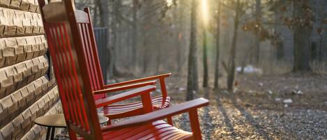 A wooden chair with a high backrest and armrests, positioned outdoors with a brick wall to the left and a forested area in the background.