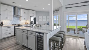 A modern kitchen with white cabinetry, a large island with stools, a view of the ocean, and a dining area with outdoor seating.