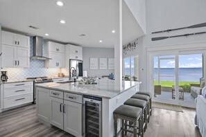 Uma cozinha moderna com armários brancos, uma ilha grande com banquetas, vista para o oceano e área de jantar com assentos externos.