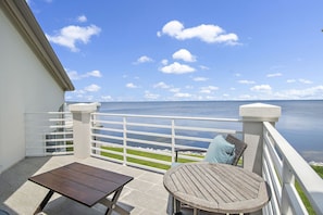 A balcony with a view of the sea, a wooden table, and a wicker chair. - 🌅 Sunset Serenity @ Sandestin | Bayfront | Golf Cart | 3BR Home (Miramar Beach)