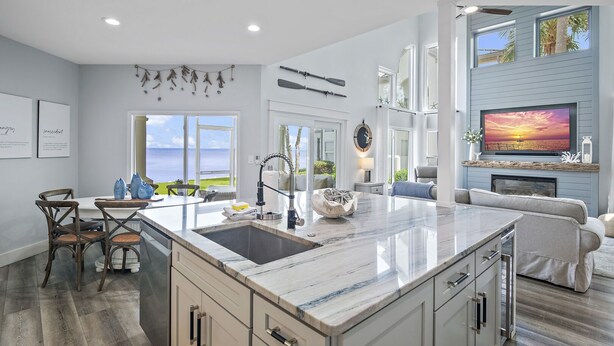 A spacious kitchen with a large island, a dining area with chairs, a living room with a sofa and TV, and large windows with a view.
