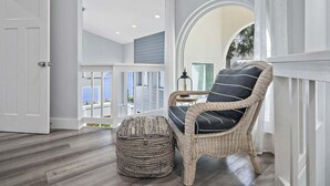 A wicker chair with a cushion, a woven ottoman, and a view of the ocean through a balcony.