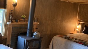 A cozy room with a wood-burning stove, a bed with a quilt, a chair, and a small shelf with decorative items. - Homestead Living at Lake Wenatchee (Leavenworth)