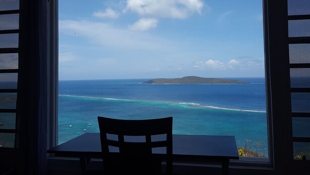 Mesa e cadeira de frente para uma vista costeira através de uma janela, com vista para o oceano e uma ilha distante.