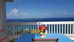 Varanda com mesa e um vaso de flores, com vista para a costa, o oceano e uma ilha distante.
