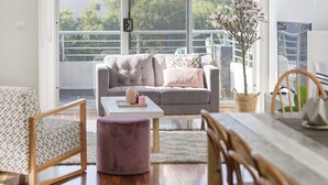 Een moderne woonkamer met een grijze bank, een klein tafeltje en een paars voetenbankje. Er is een balkon met een hekwerk en uitzicht op bomen.