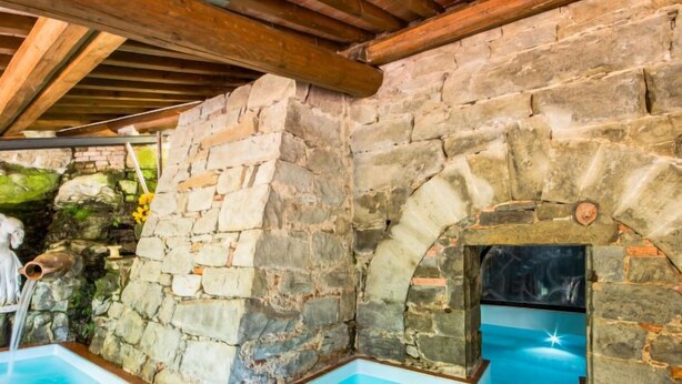 Un espace piscine aux murs de pierre, doté d’un plafond en bois rustique et d’une arche en pierre menant à un intérieur sombre.