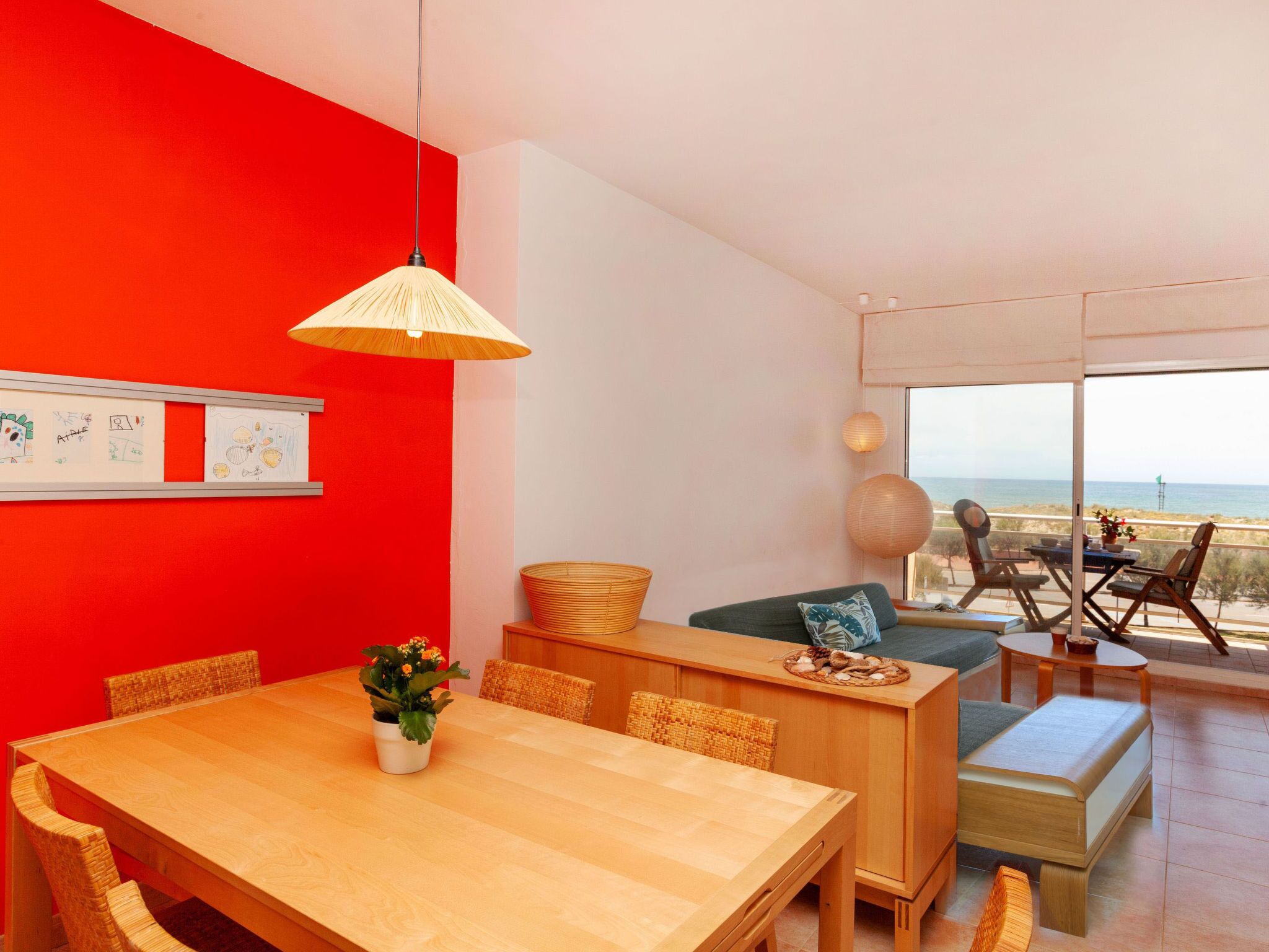 En moderne stue med en rød accentvæg, et træspisebord, fletstole, en sofa og en balkon med havudsigt.