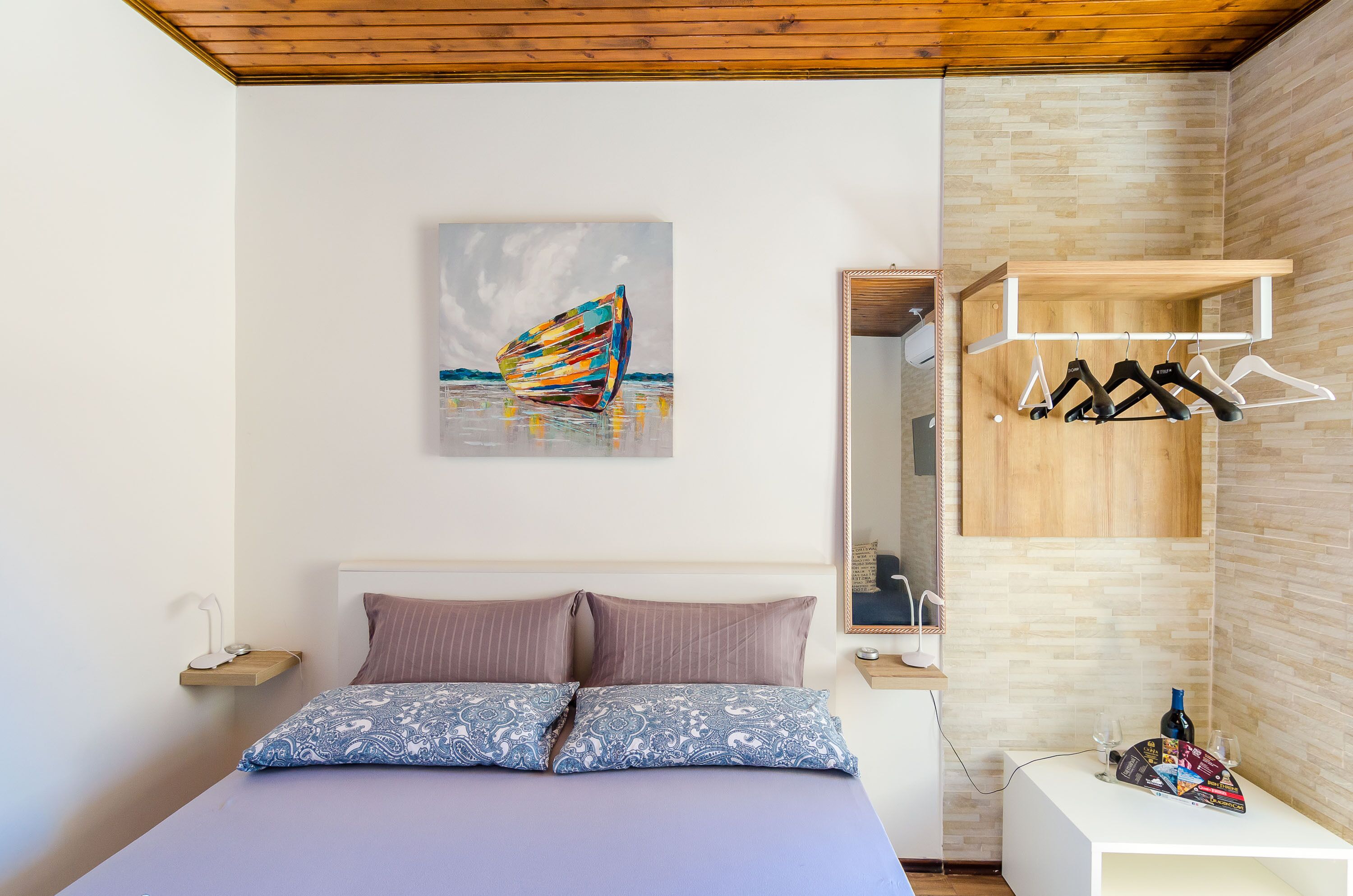 Ein Schlafzimmer mit Bett, Kissen, einem Gemälde, einer Holzablage mit Kleidung und einem kleinen Tisch mit Gläsern und einer Flasche.