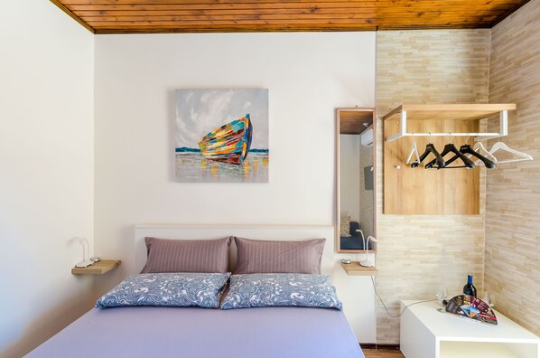 A bedroom with a bed, pillows, a painting, a wooden shelf with clothes, and a small table with glasses and a bottle.