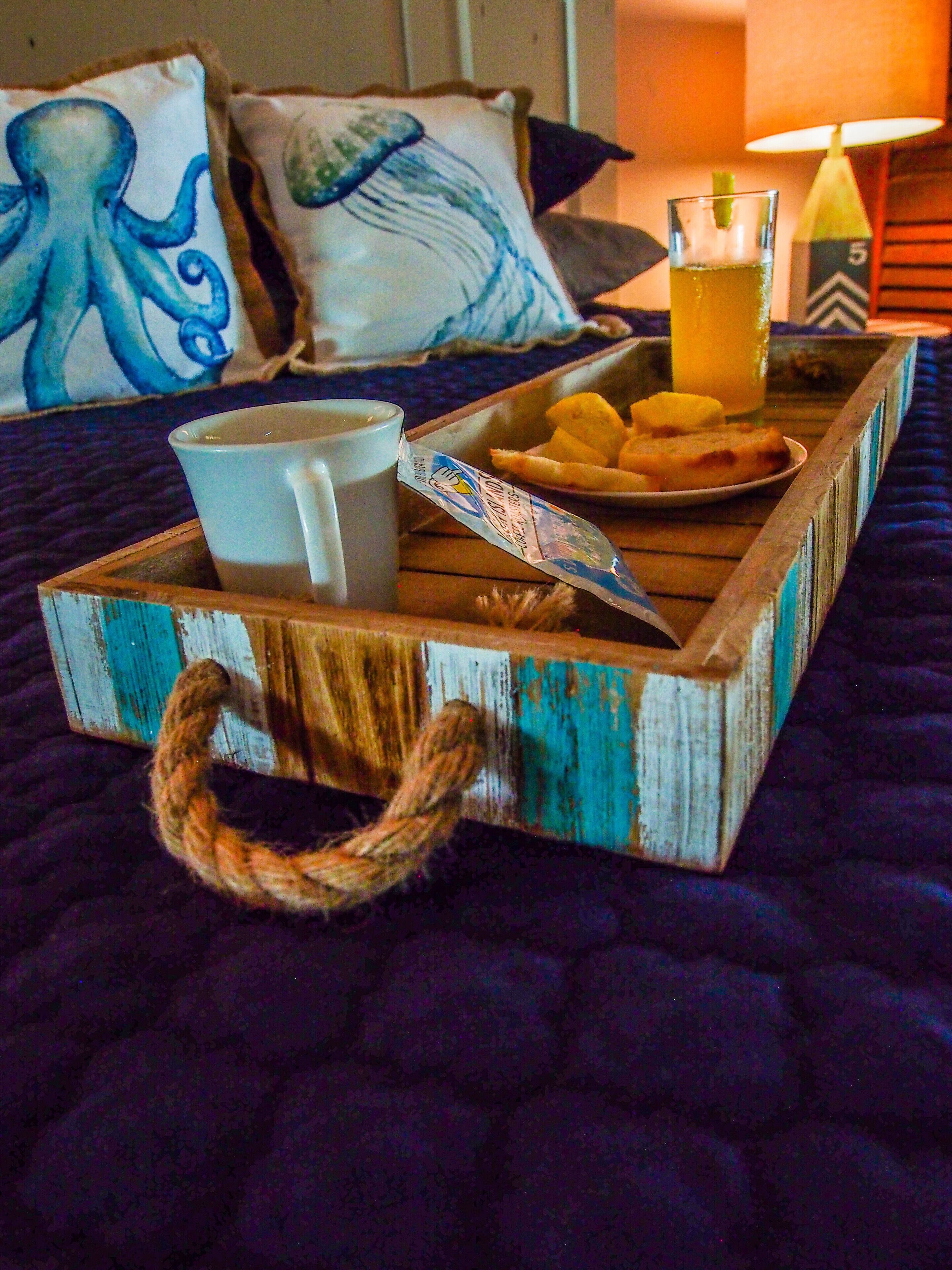 Ein Holztablett mit einer Tasse, einem Glas Saft und einem Teller Essen auf einem Bett mit einer blauen Bettdecke.