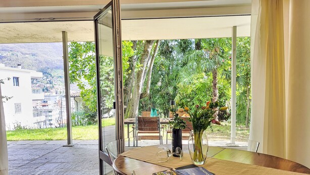 Un coin repas avec une table dressée pour deux personnes, un vase rempli de fleurs et une vue sur un jardin luxuriant et des bâtiments éloignés.